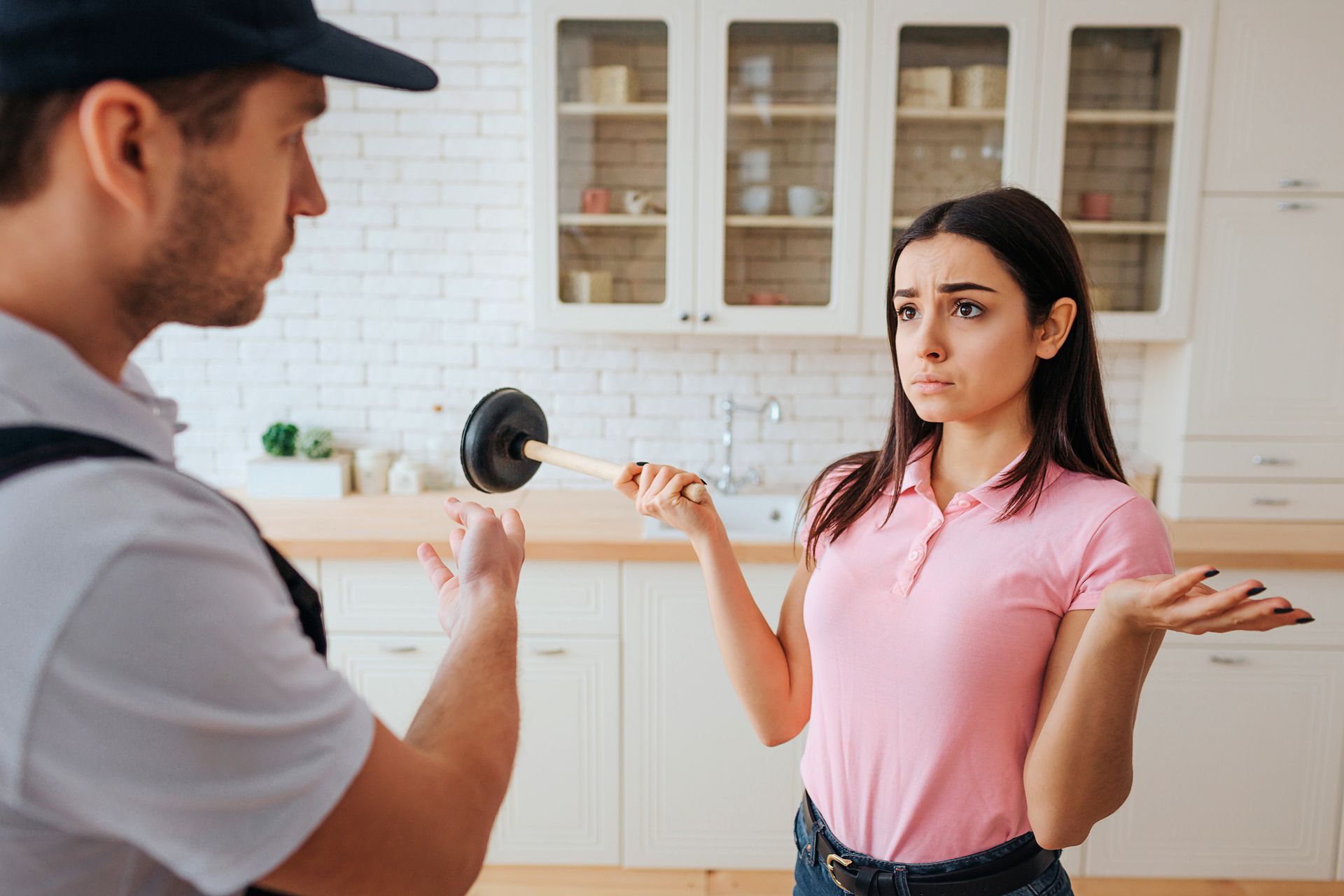 Woman with plunger consulting plumber in modern kitchen about clogged sink plumbing issue.