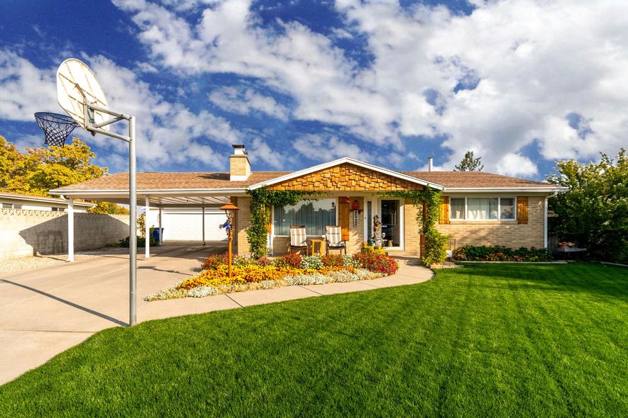 House with carport, basketball hoop, and vibrant flower bed under a cloudy blue sky.