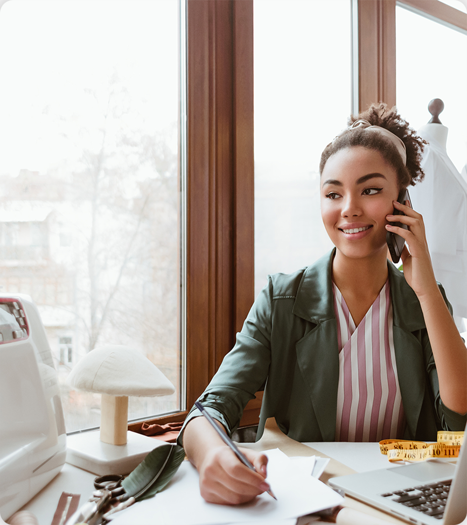 Woman in green jacket talking on phone, writing at desk near window.