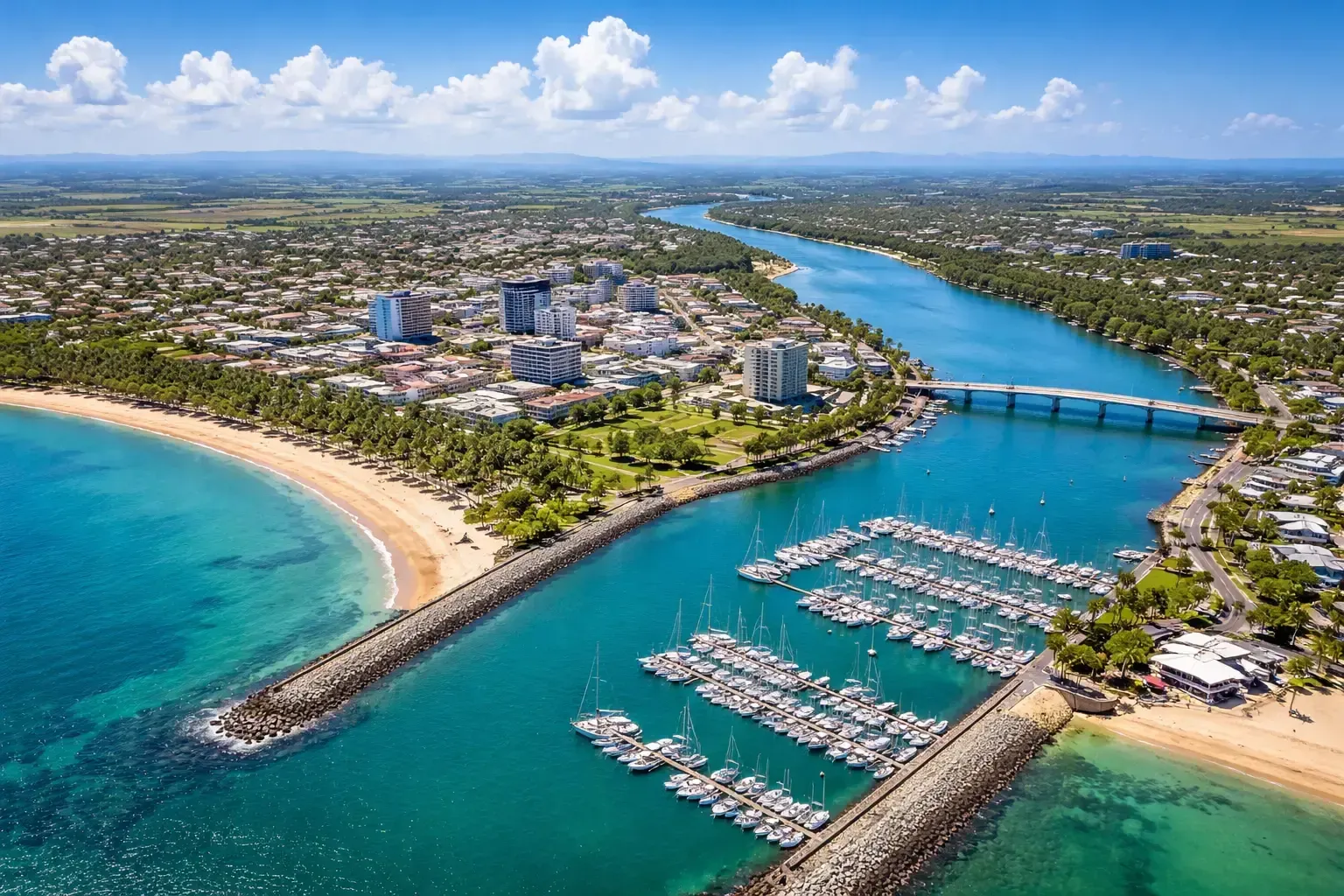 Aerial view of a Mackay Marina with a river flowing into the ocean, marina, and beach on a sunny day.