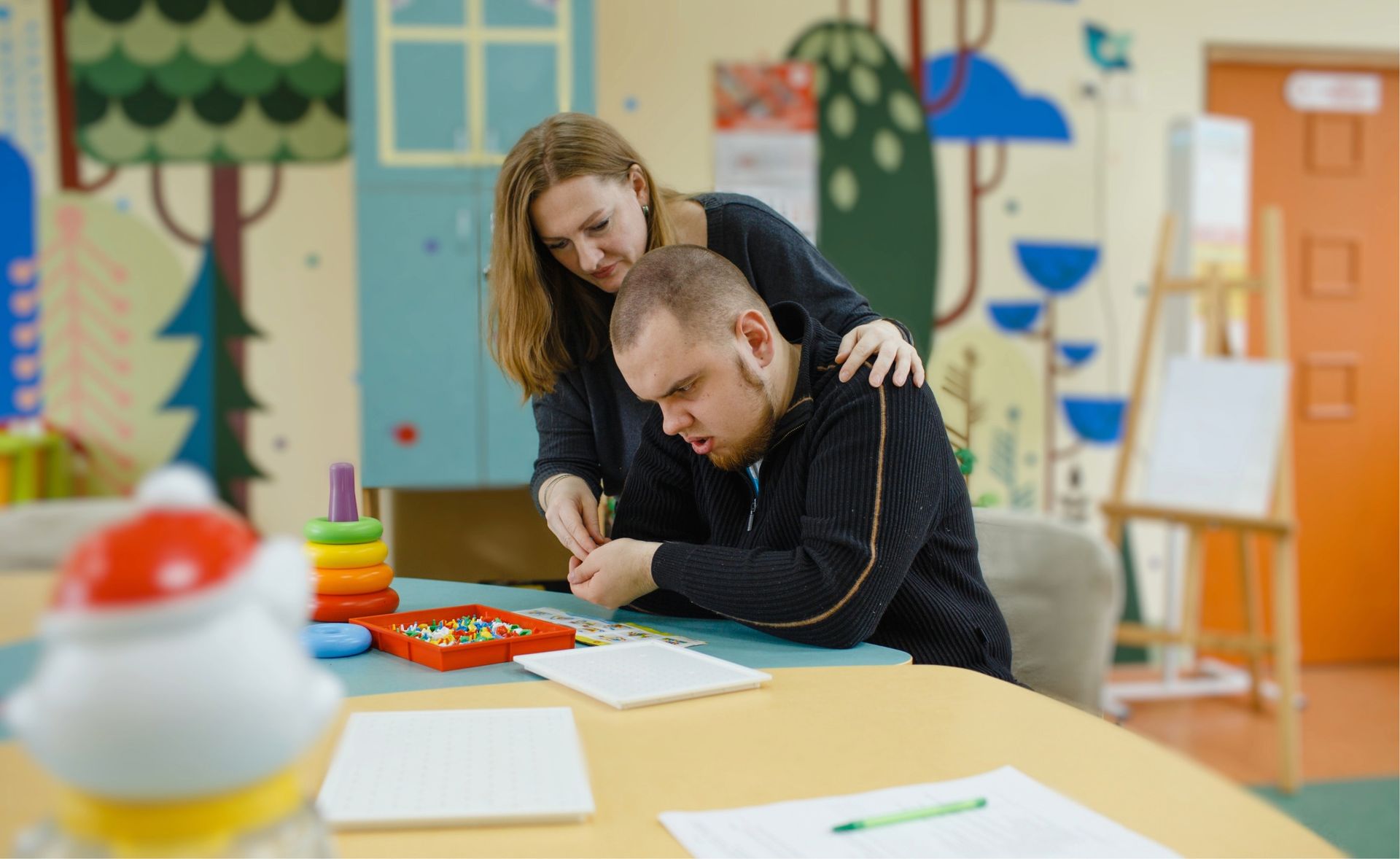 Woman assisting a man with crafting; they sit at a table in a brightly decorated room.