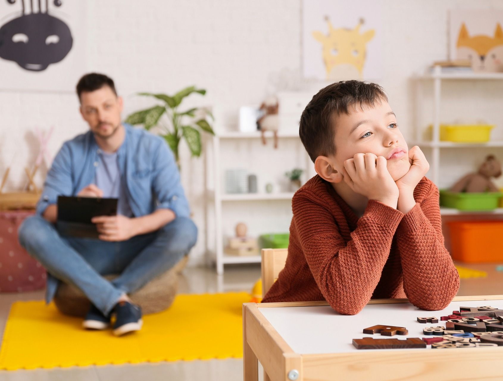 Boy resting chin on hands at table, puzzle pieces scattered. Man with tablet sits nearby. Bright room.