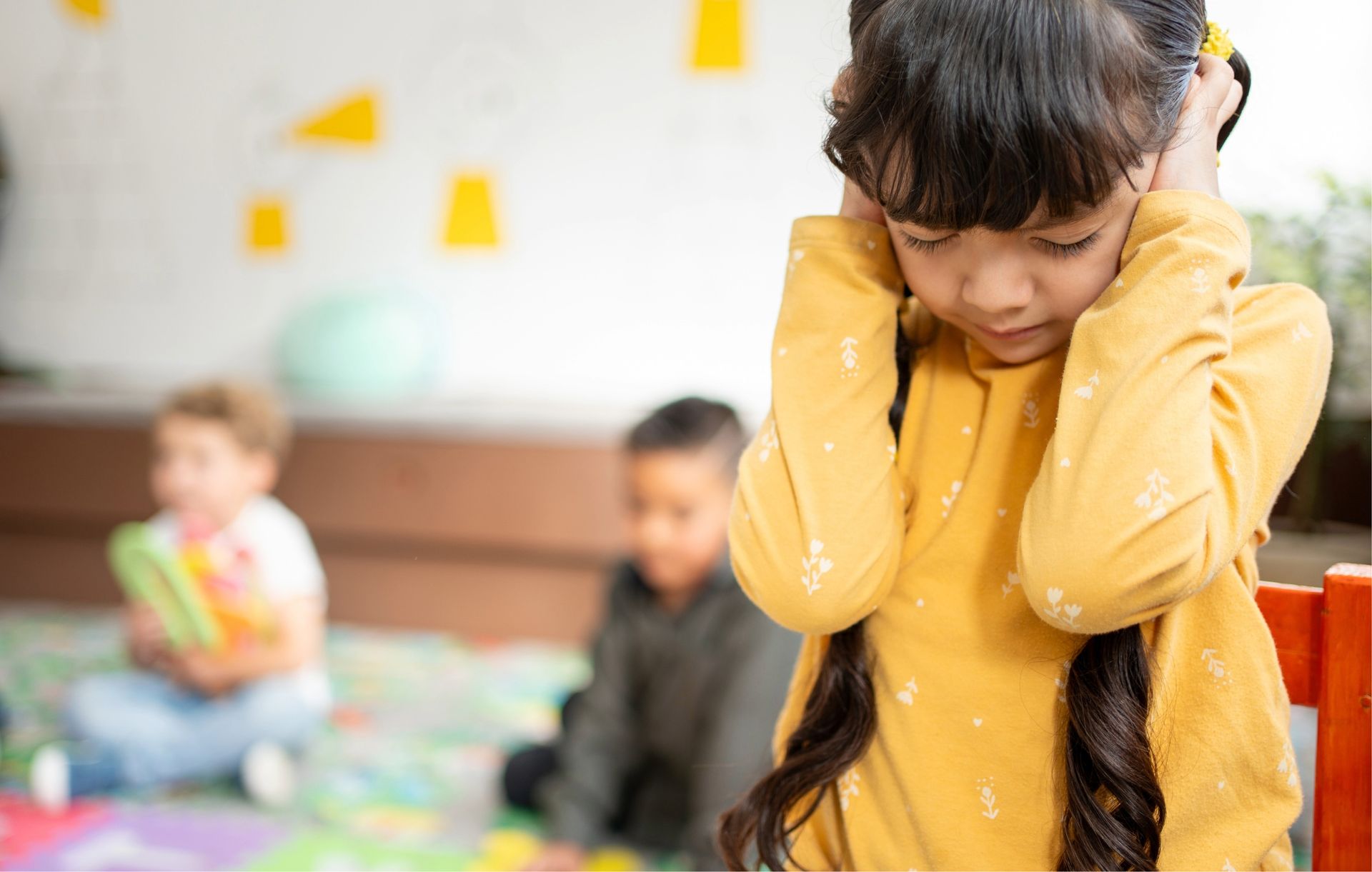 Girl with long braids covers ears, looking down, in a classroom with other children sitting.