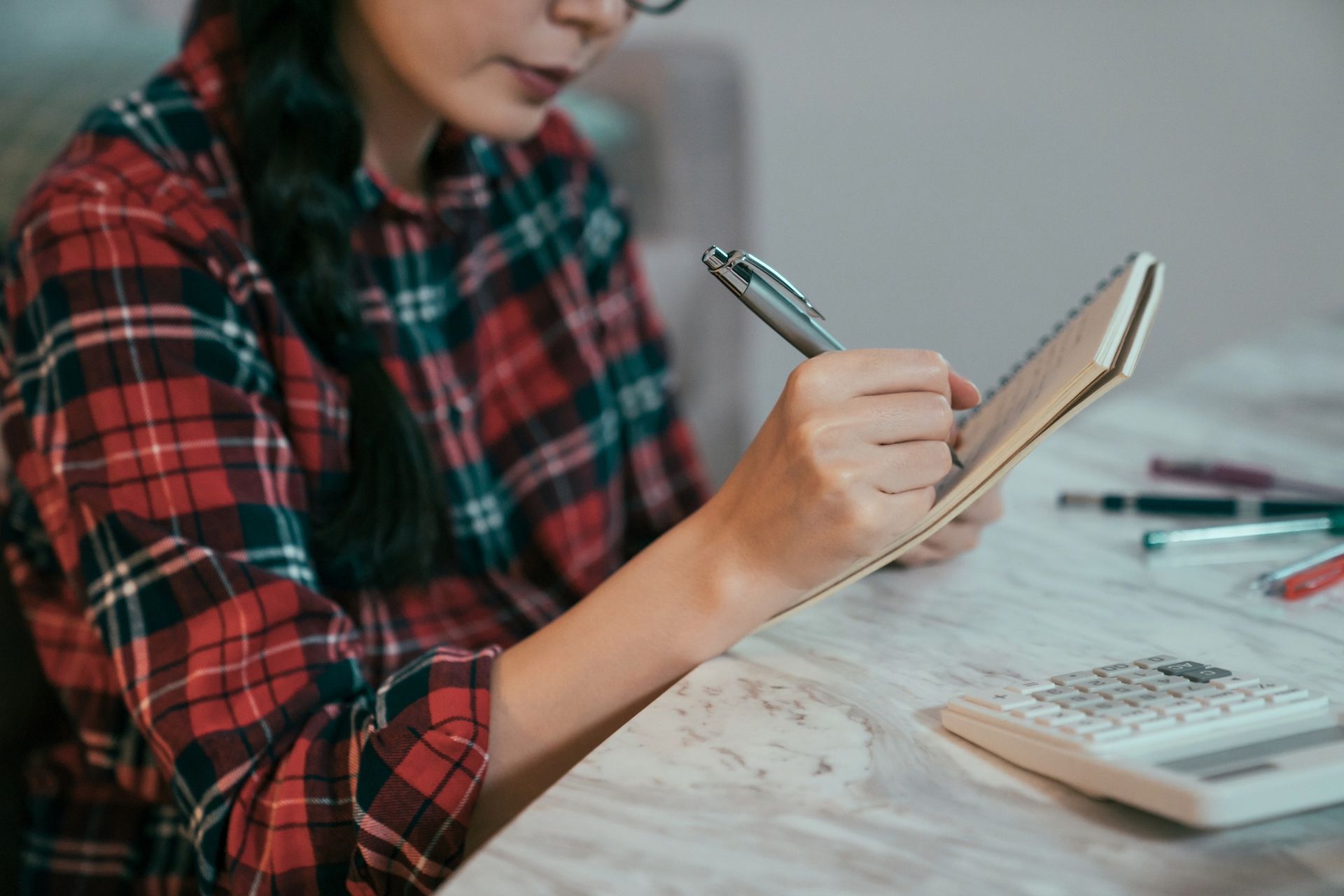 Person in plaid shirt writing in notebook with a pen at a table; calculator visible.