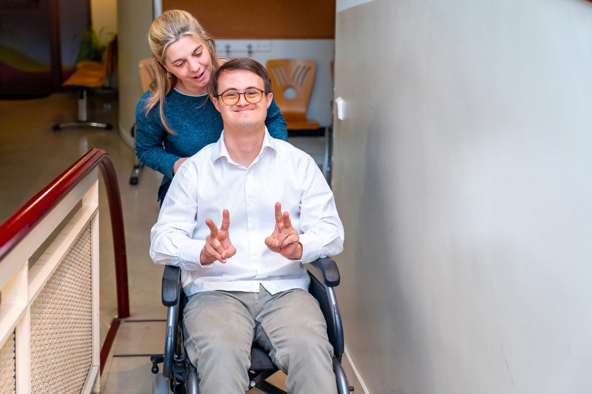Woman assists a person in a wheelchair on a ramp. The person in the wheelchair makes a peace sign.