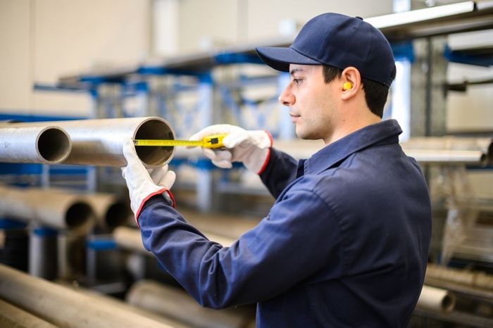 A man is measuring a pipe with a tape measure in a factory.