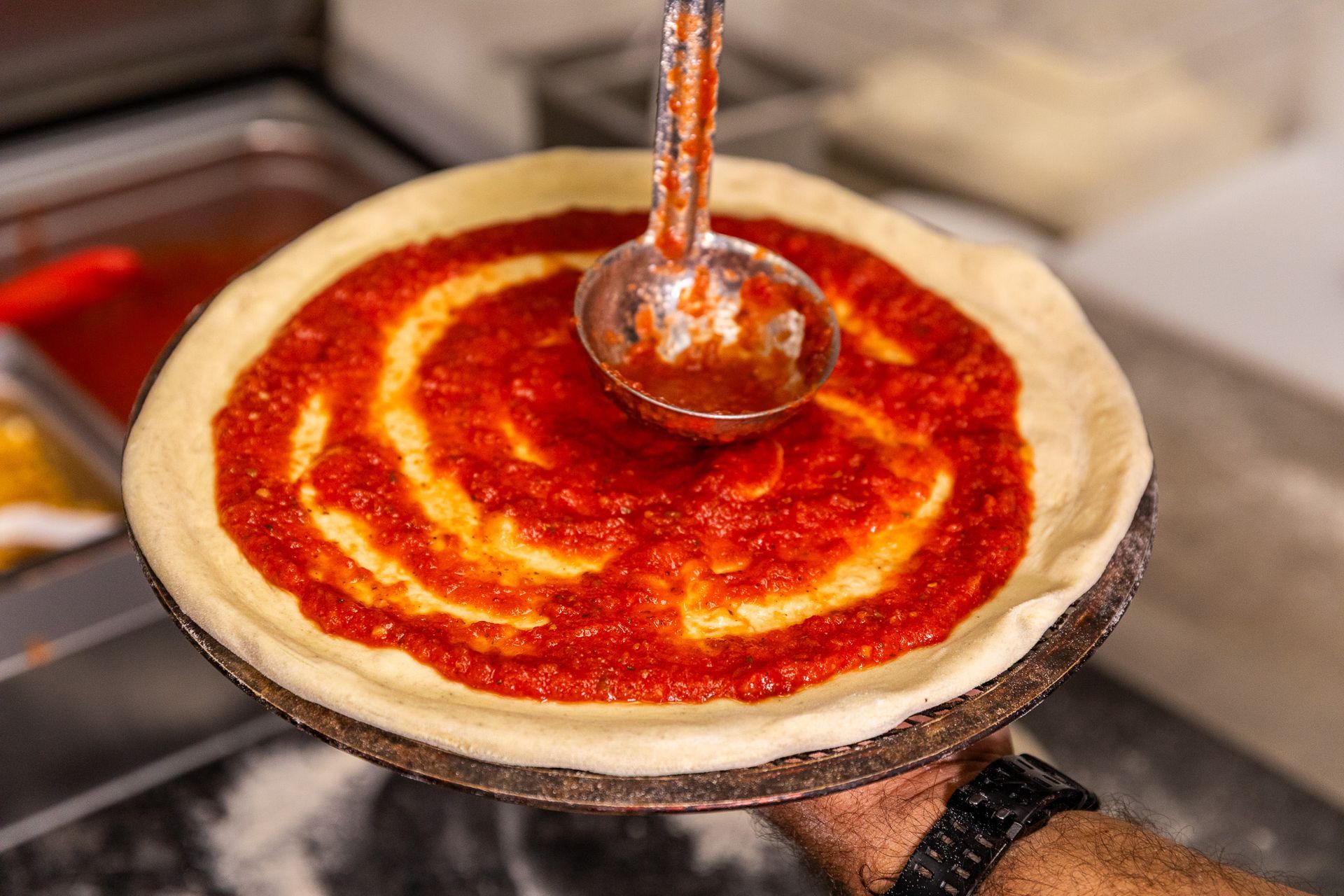 A person is spreading tomato sauce on a pizza with a ladle.