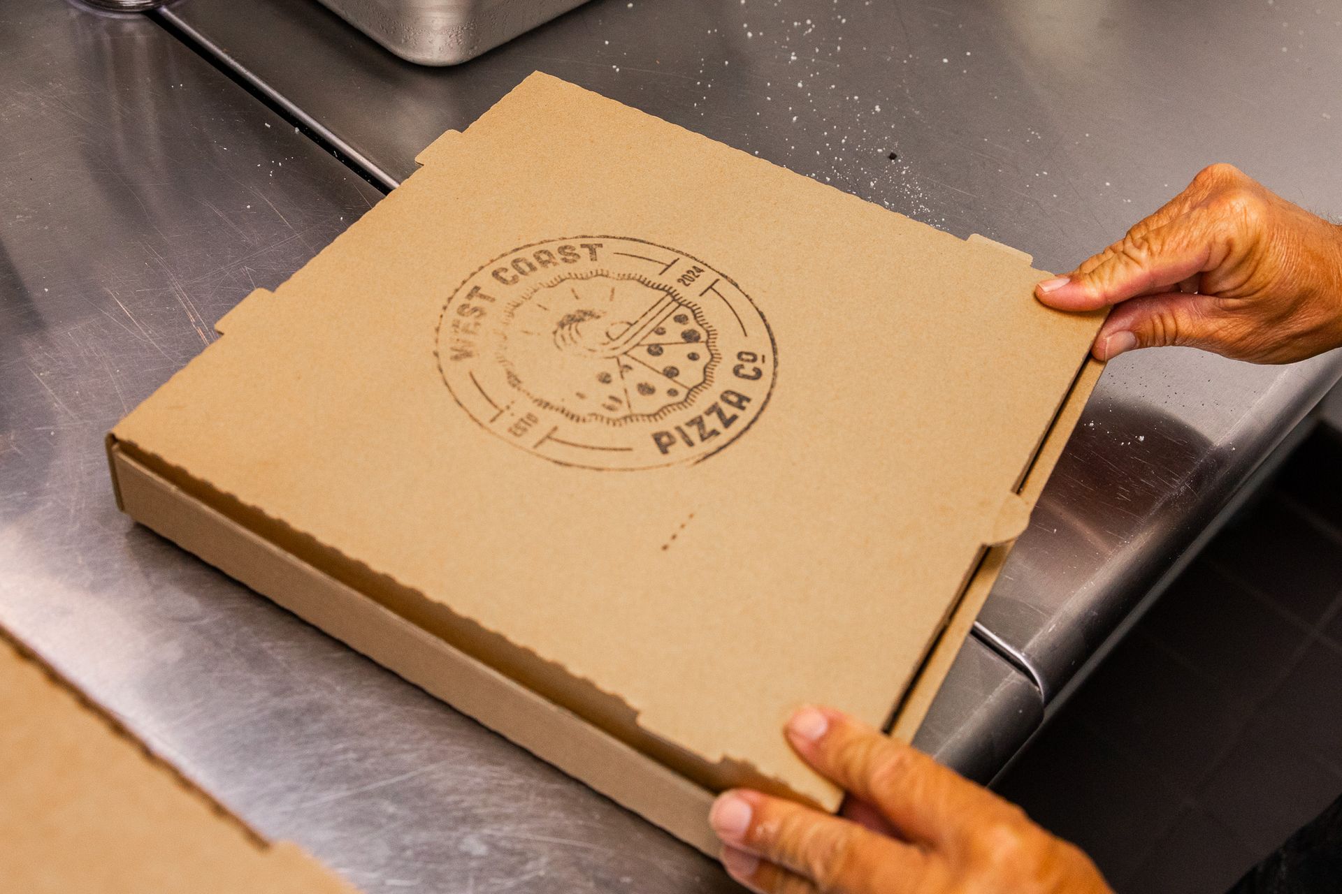 A person is opening a pizza box on a stainless steel counter.