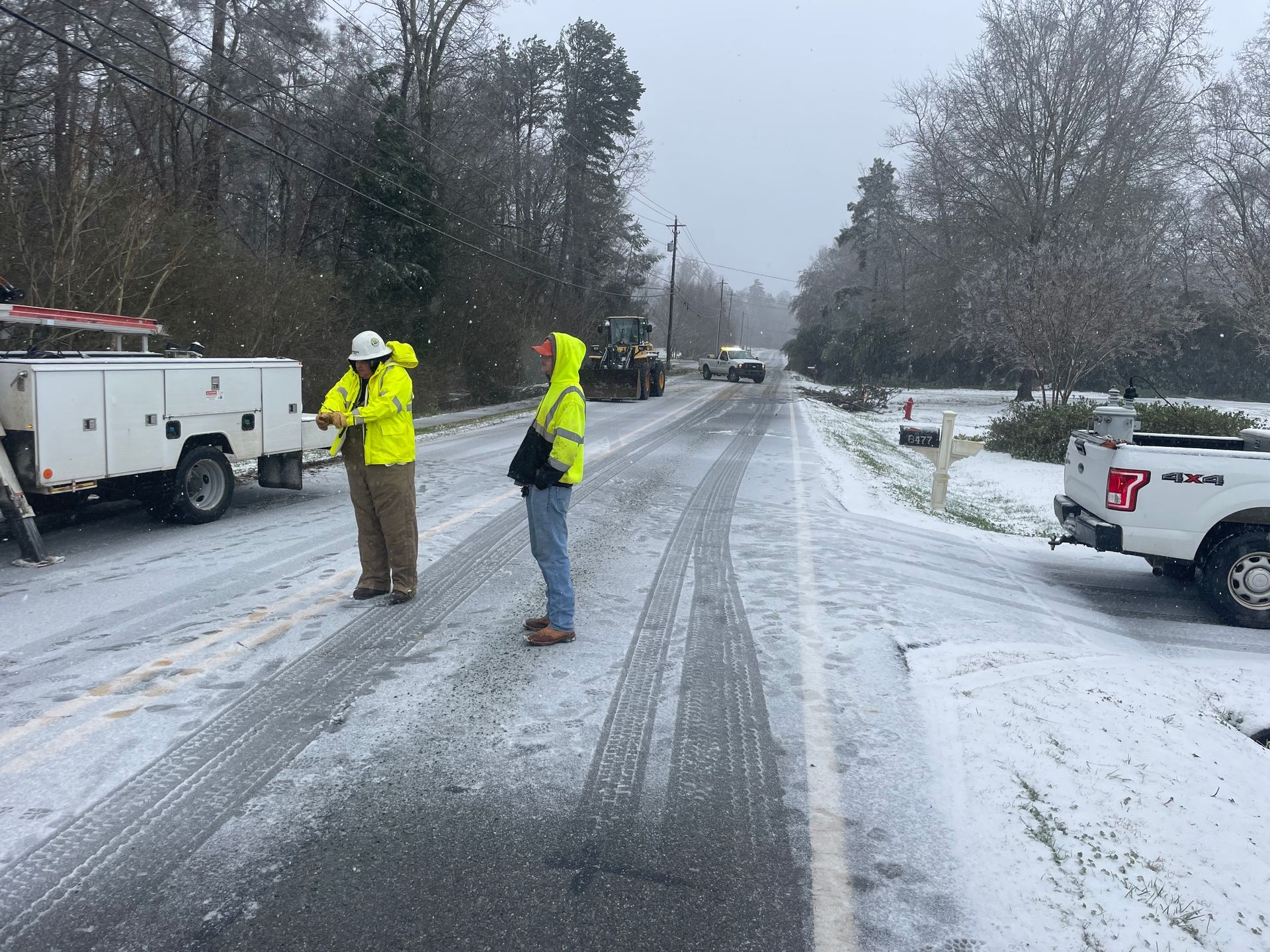 Snowy road with utility workers near trucks, wearing bright yellow safety vests.