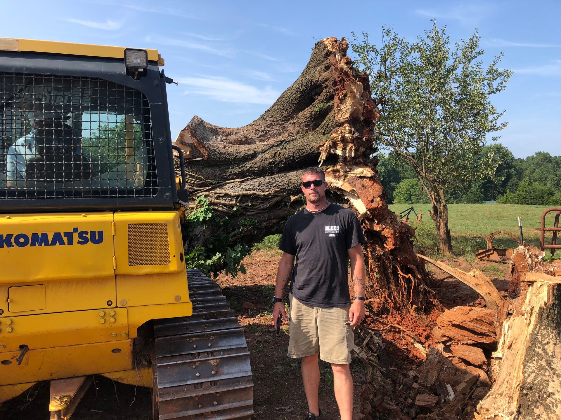 Man stands next to a yellow Komatsu bulldozer and large fallen tree, outdoors.