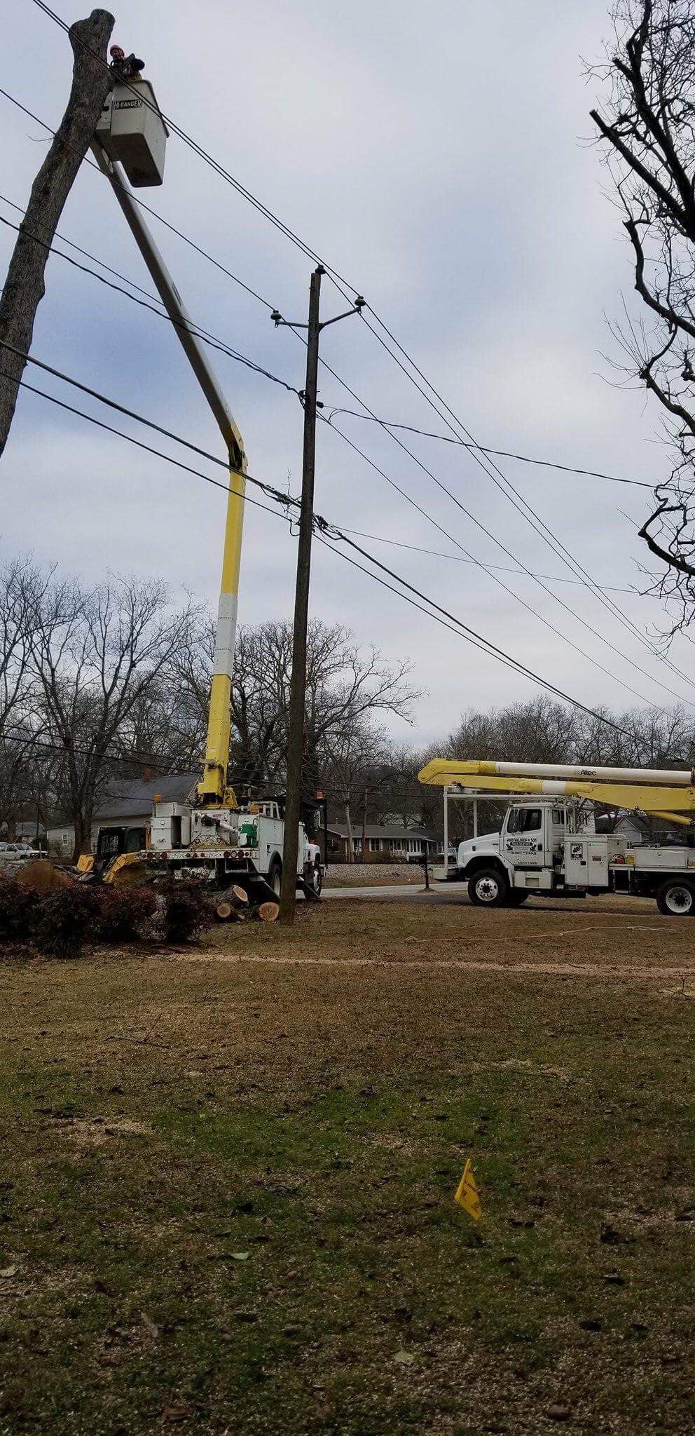 Utility trucks with raised booms repairing power lines. Cloudy sky, trees, and grass in the foreground.