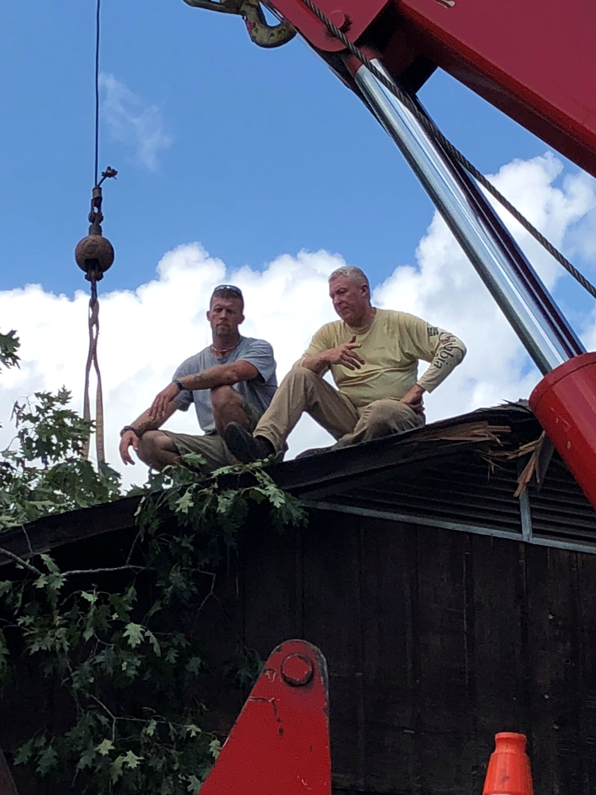 Two men squat on a damaged roof. A crane and trees are visible against a cloudy sky.