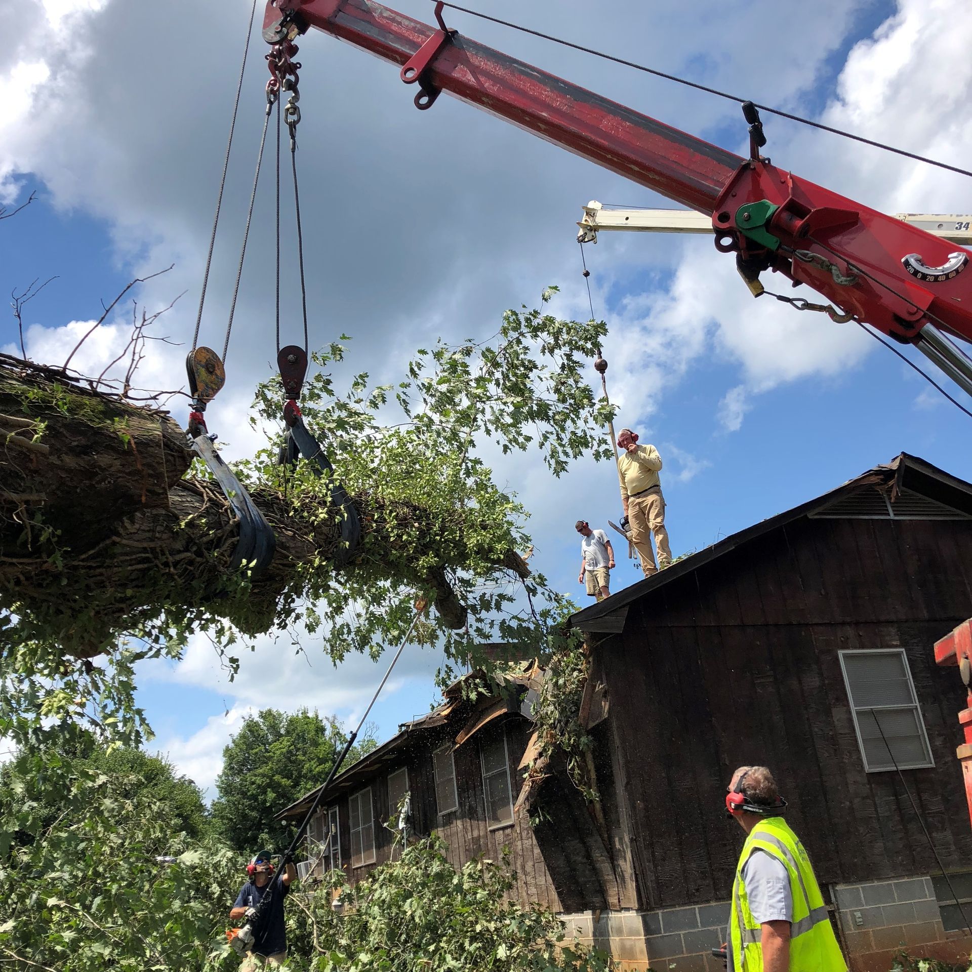 Crane lifting tree branches from a damaged barn roof. Workers in safety gear oversee.