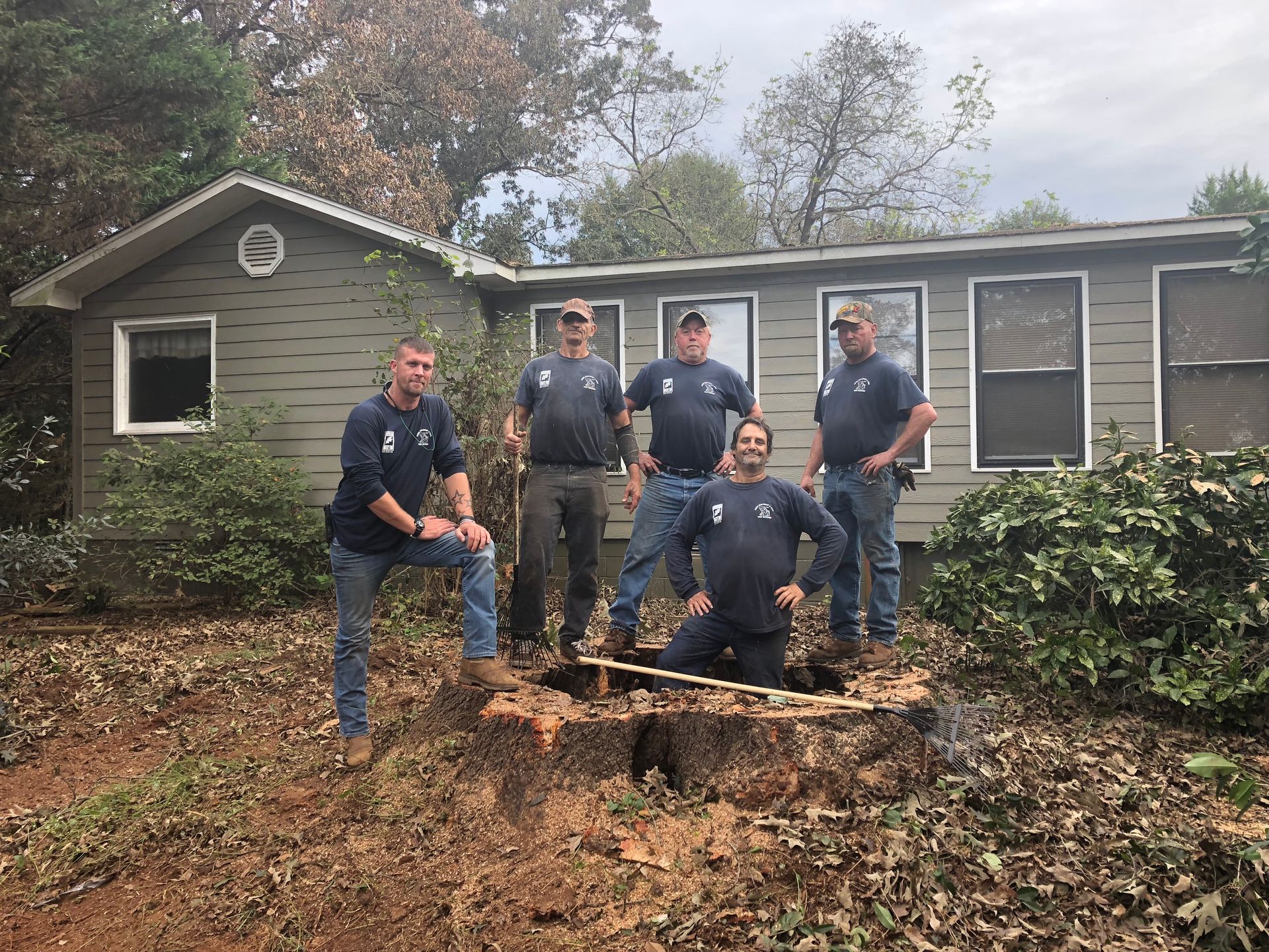 Five men in work clothes pose near a tree stump in front of a house.