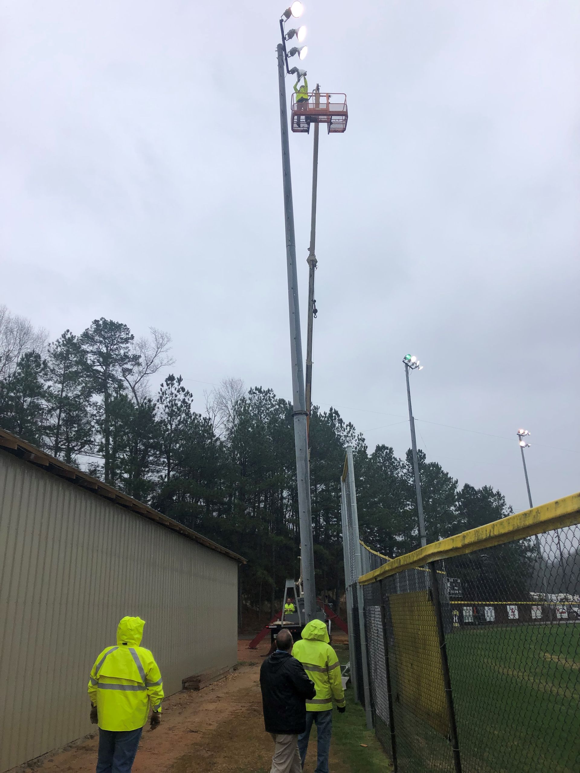 Workers in neon vests adjust stadium lights from a lift, near a field with a fence, on a cloudy day.