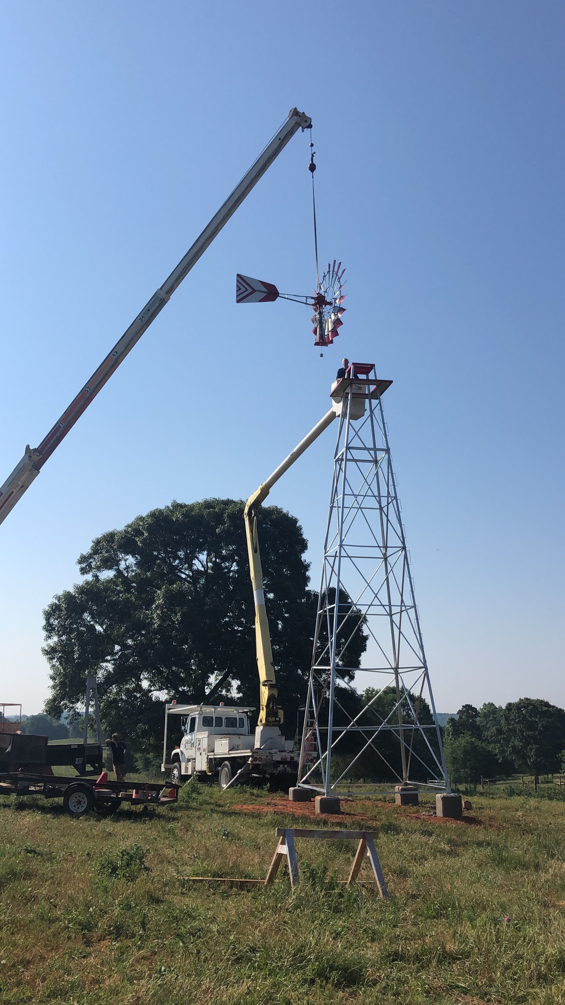 Cranes installing a windmill atop a tall, metal tower in a field under a clear blue sky.
