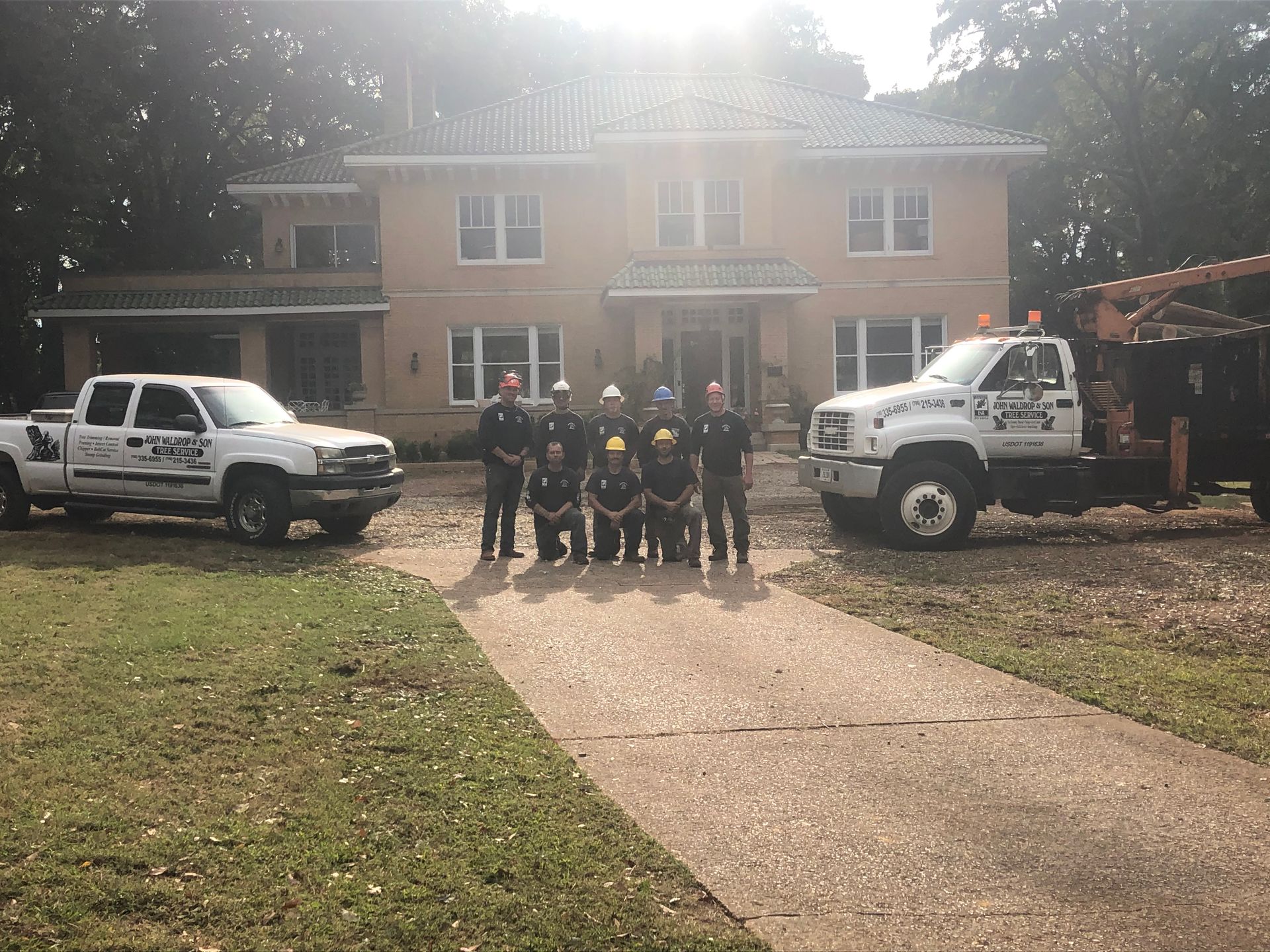 A team of tree service workers poses in front of a large house with their trucks on the driveway.