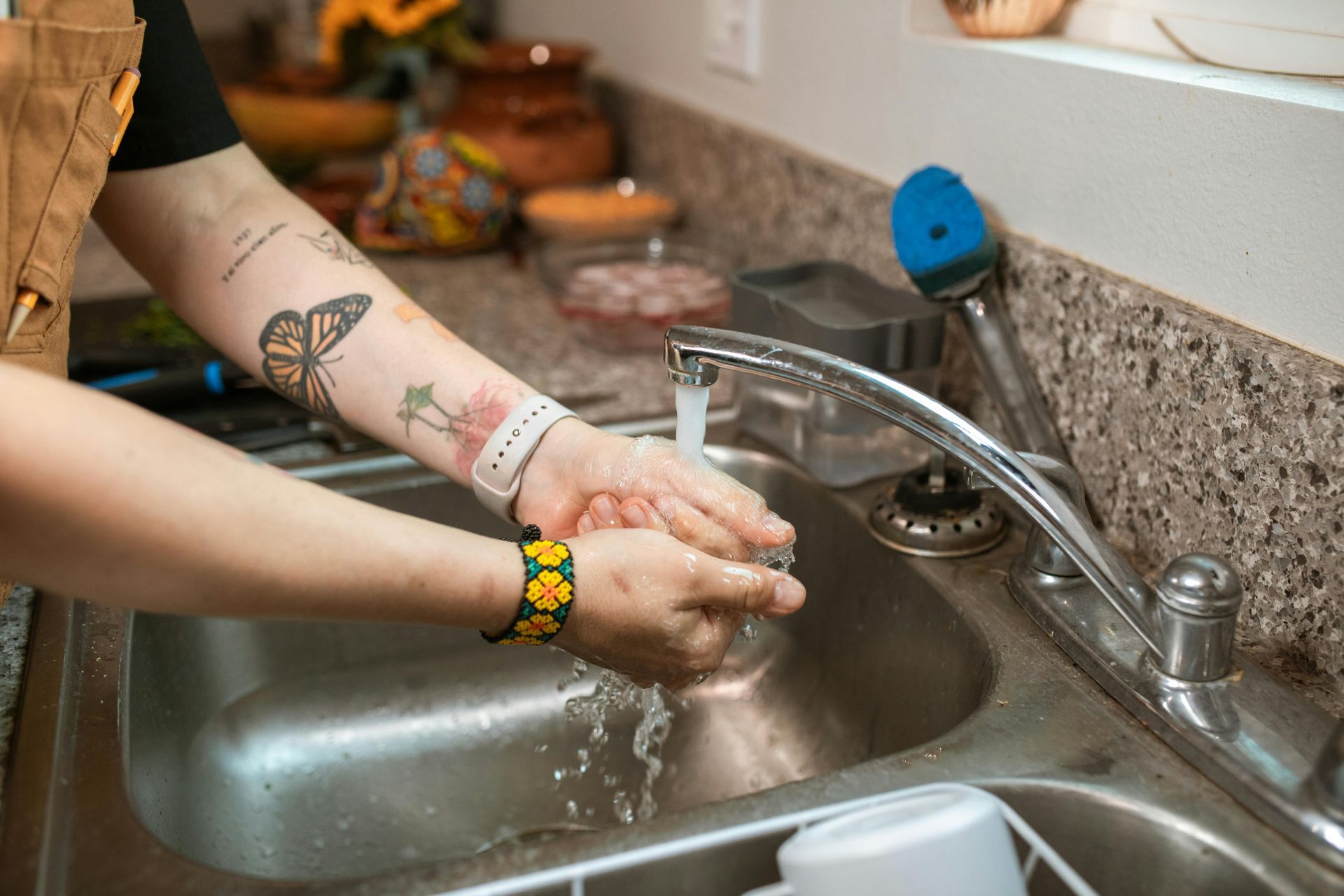 Person washes hands in a stainless steel kitchen sink, faucet running. Arm tattoos visible.
