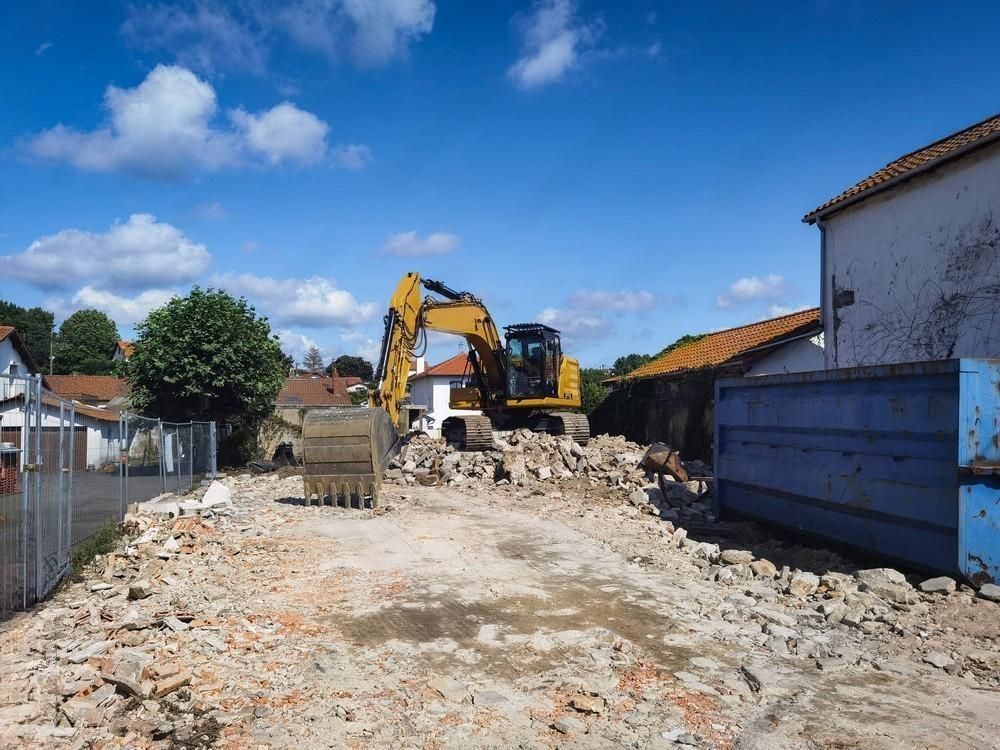 a yellow excavator is demolishing a building with a blue dumpster in the foreground