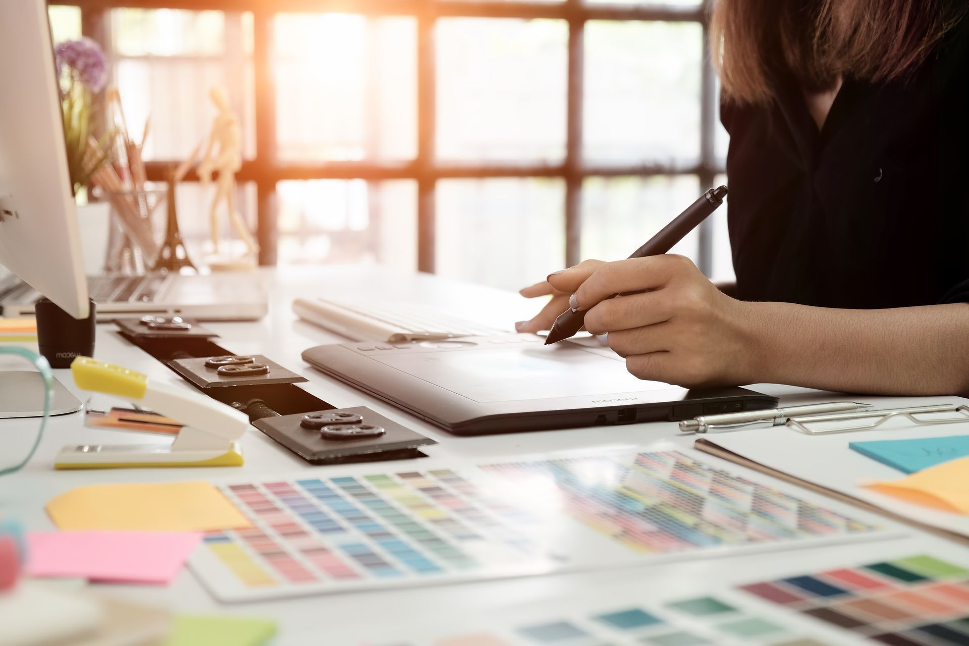 Woman using a stylus on a graphics tablet, with color swatches and office supplies on a bright desk.