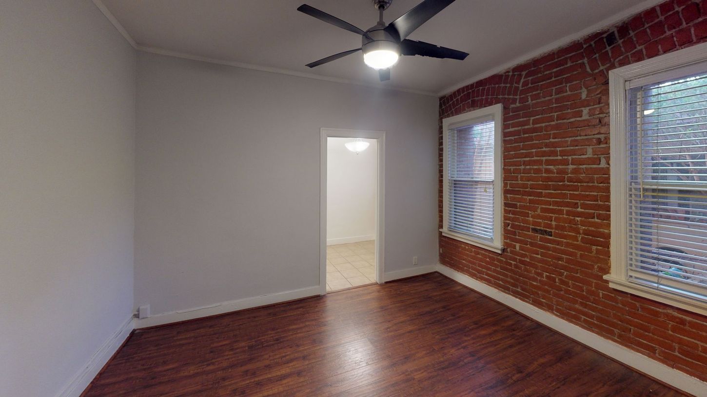 Room with exposed brick wall, windows, wooden floor, and a ceiling fan.