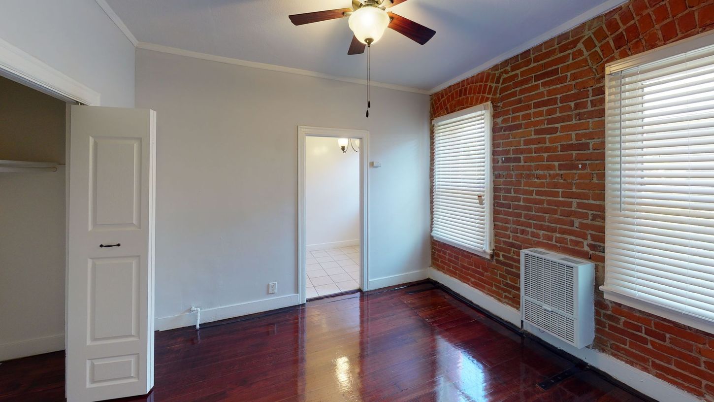 Bedroom with brick accent wall, hardwood floor, white door, and windows with blinds.