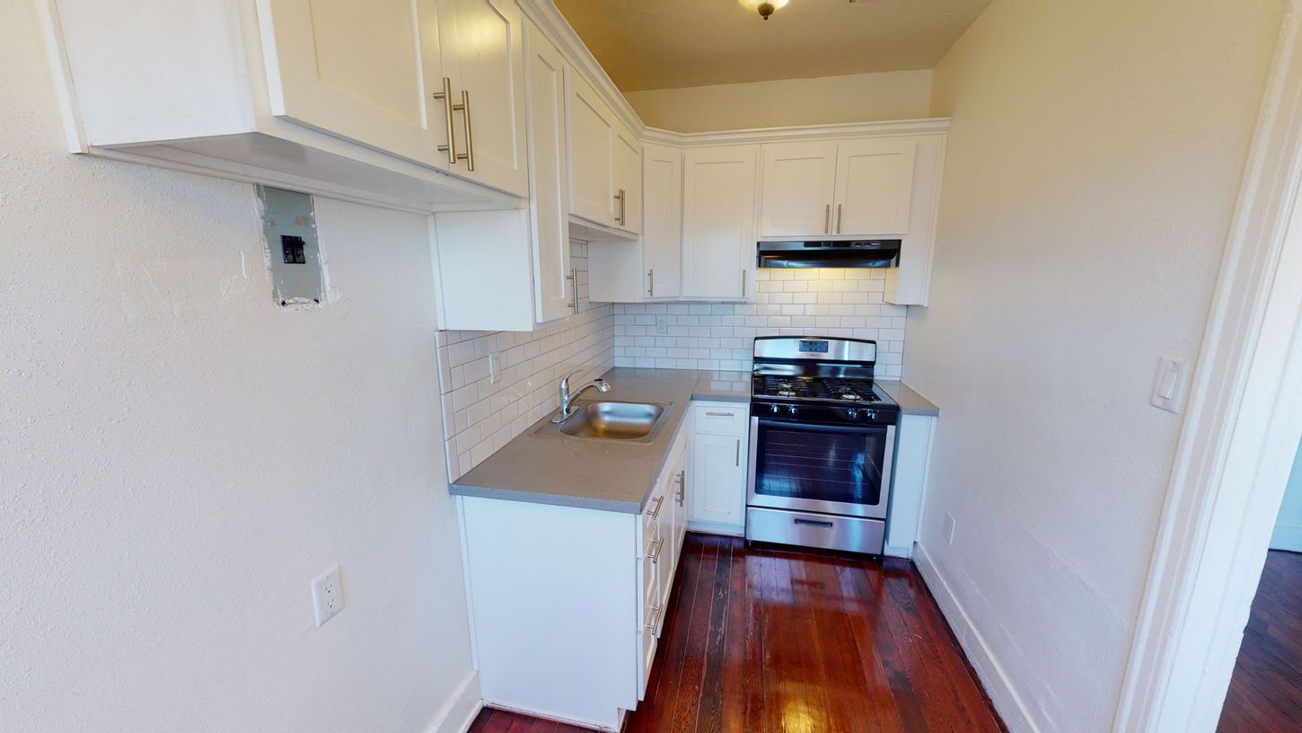 White kitchen with stainless steel appliances, cabinets, and light gray countertops. Dark wood floors.
