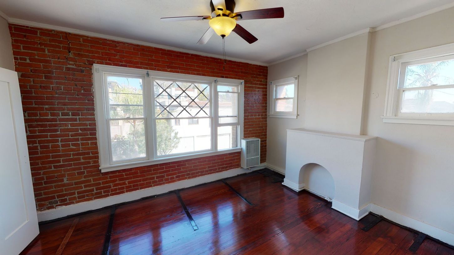 Room with exposed brick wall, windows, fireplace, dark wood floor, and ceiling fan.