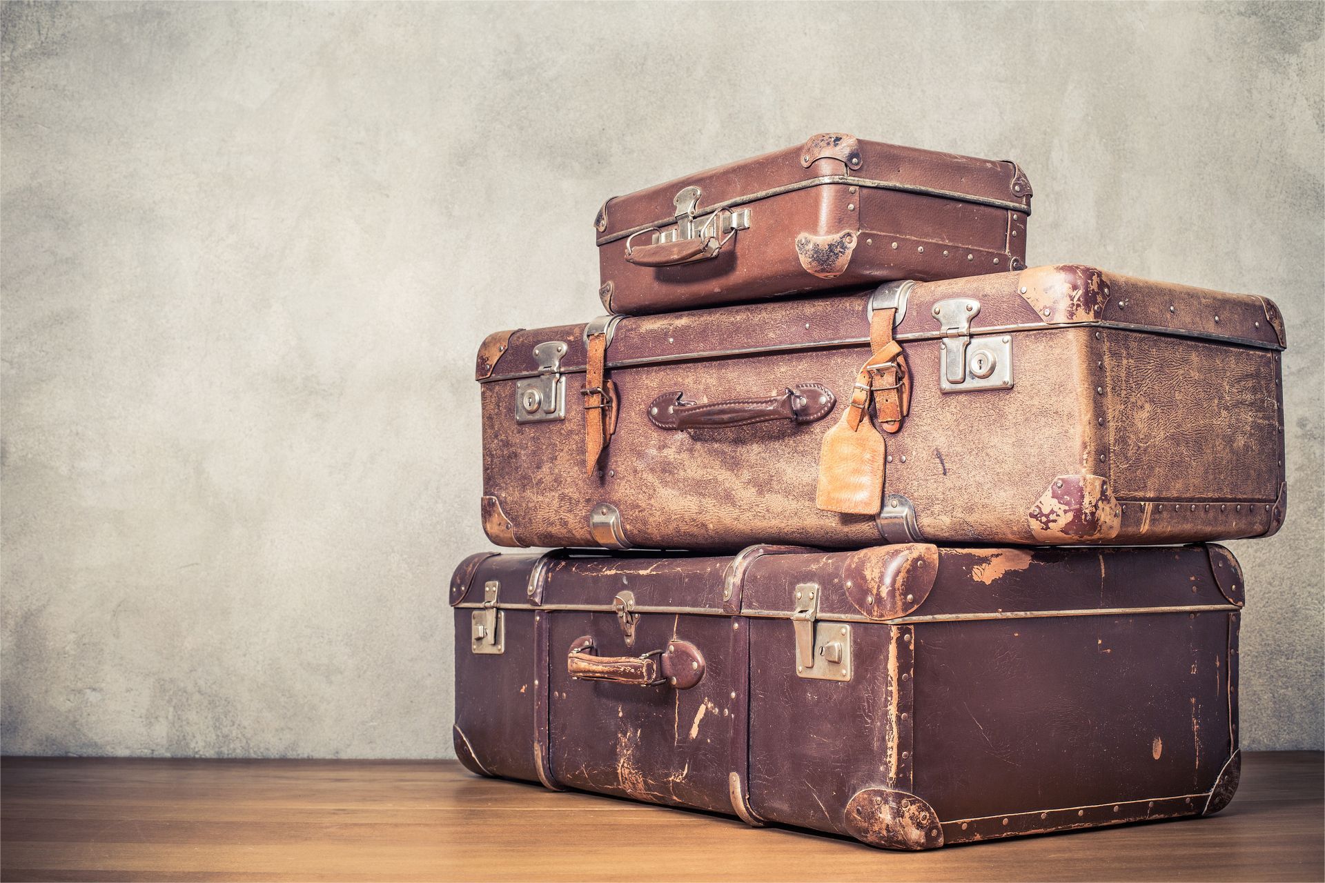 Three stacked vintage brown leather suitcases sit on a wooden surface against a neutral, textured wall.