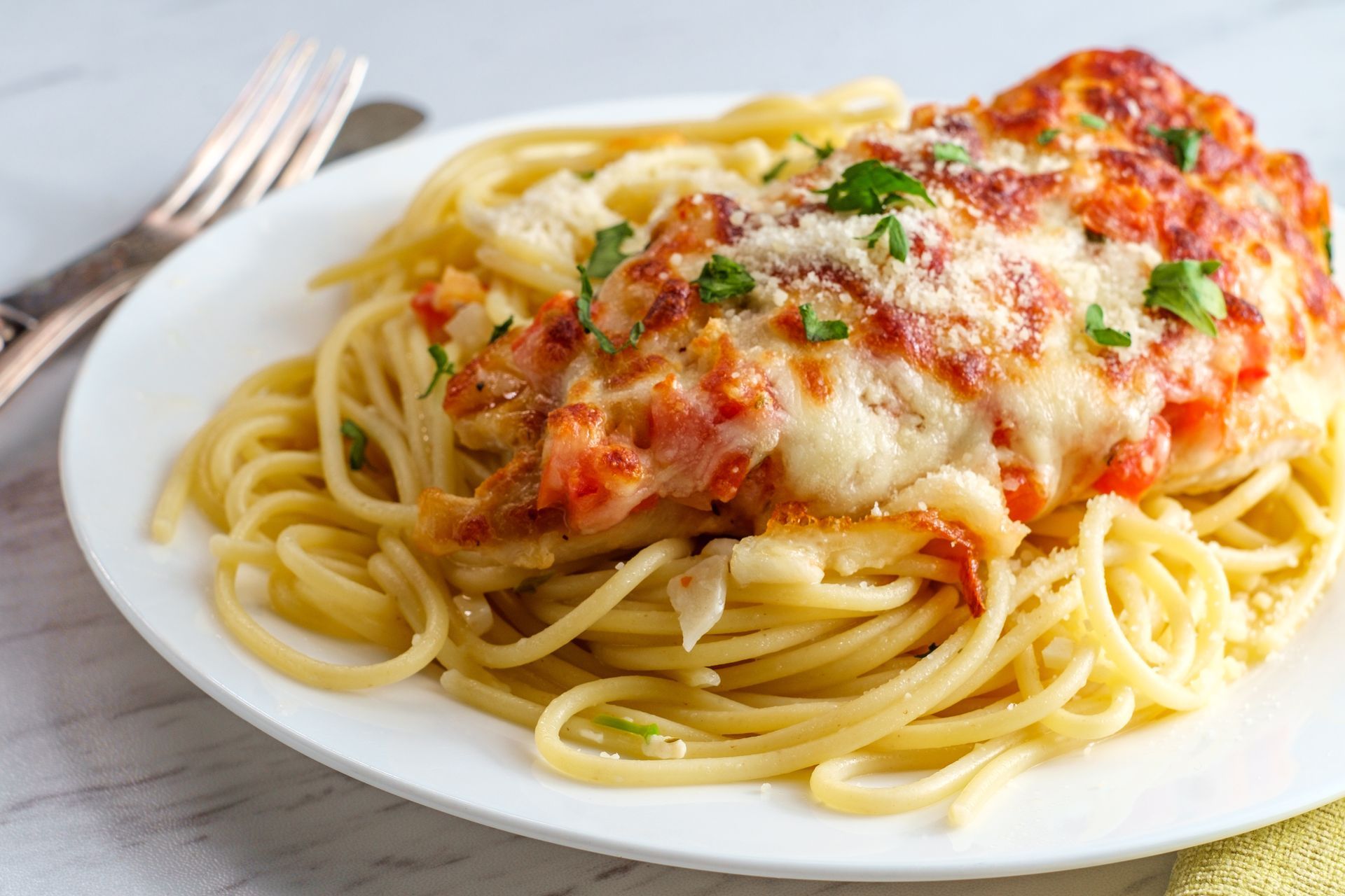 Chicken parmesan with melted cheese and red sauce served over a bed of spaghetti on a white plate with a fork nearby.