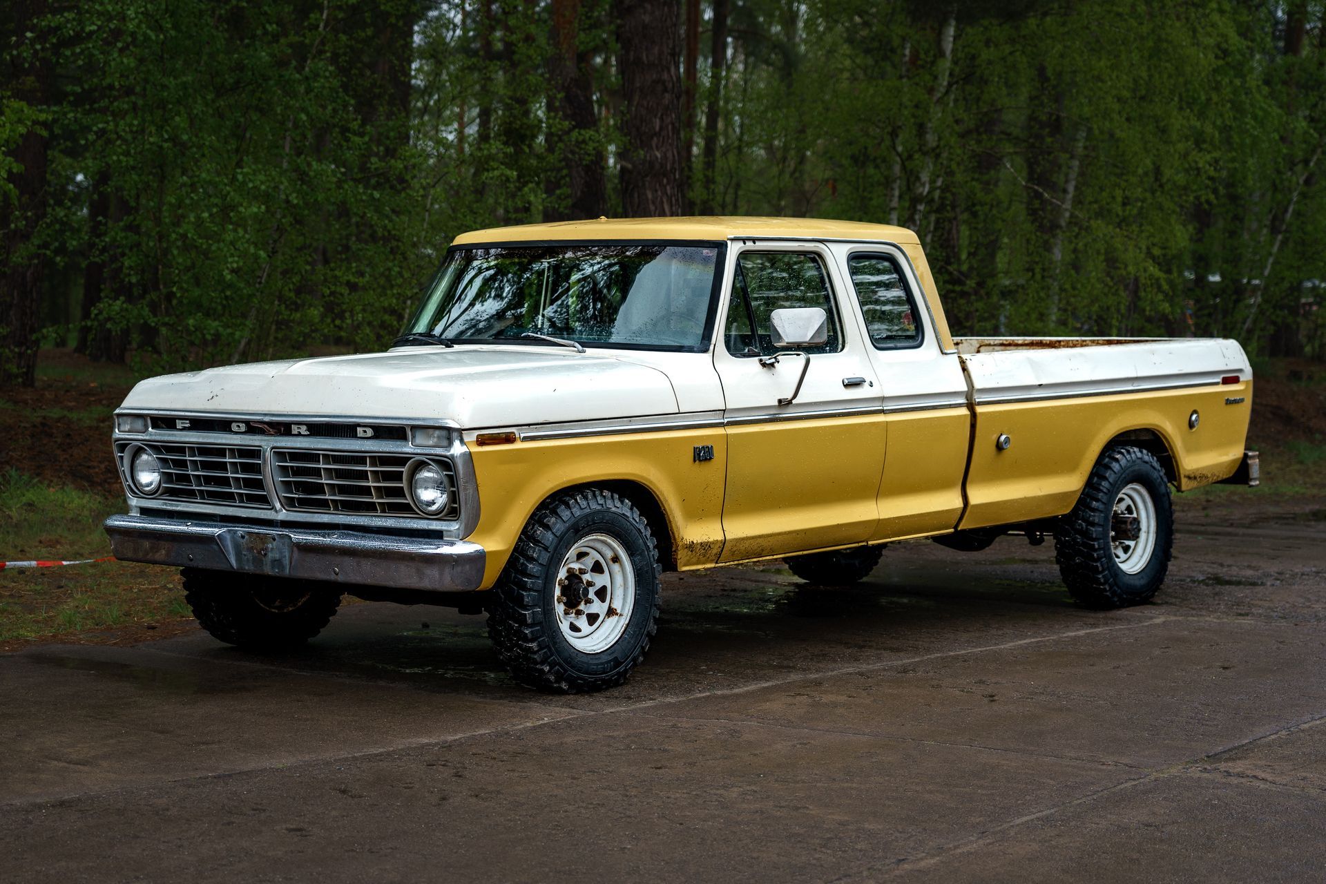 A yellow and white vintage Ford pickup truck parked on an asphalt road in front of a forest.