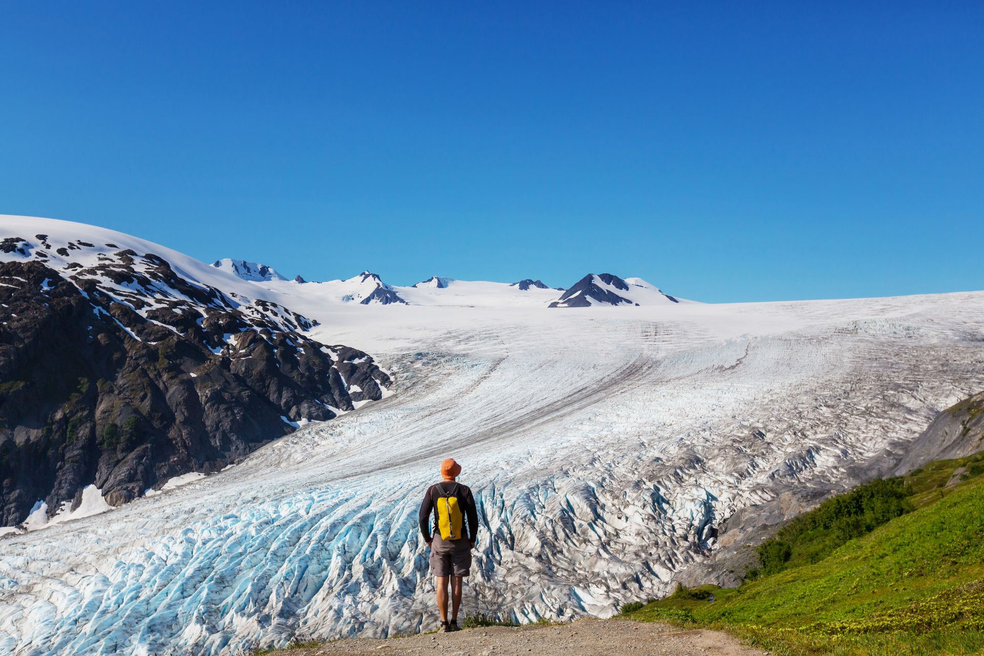 A person with a yellow backpack hikes toward a massive, blue-tinted glacier under a clear blue sky.