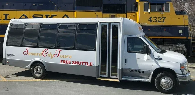 A white shuttle bus for Seward City Tours parked in front of a yellow and blue Alaska Railroad locomotive.