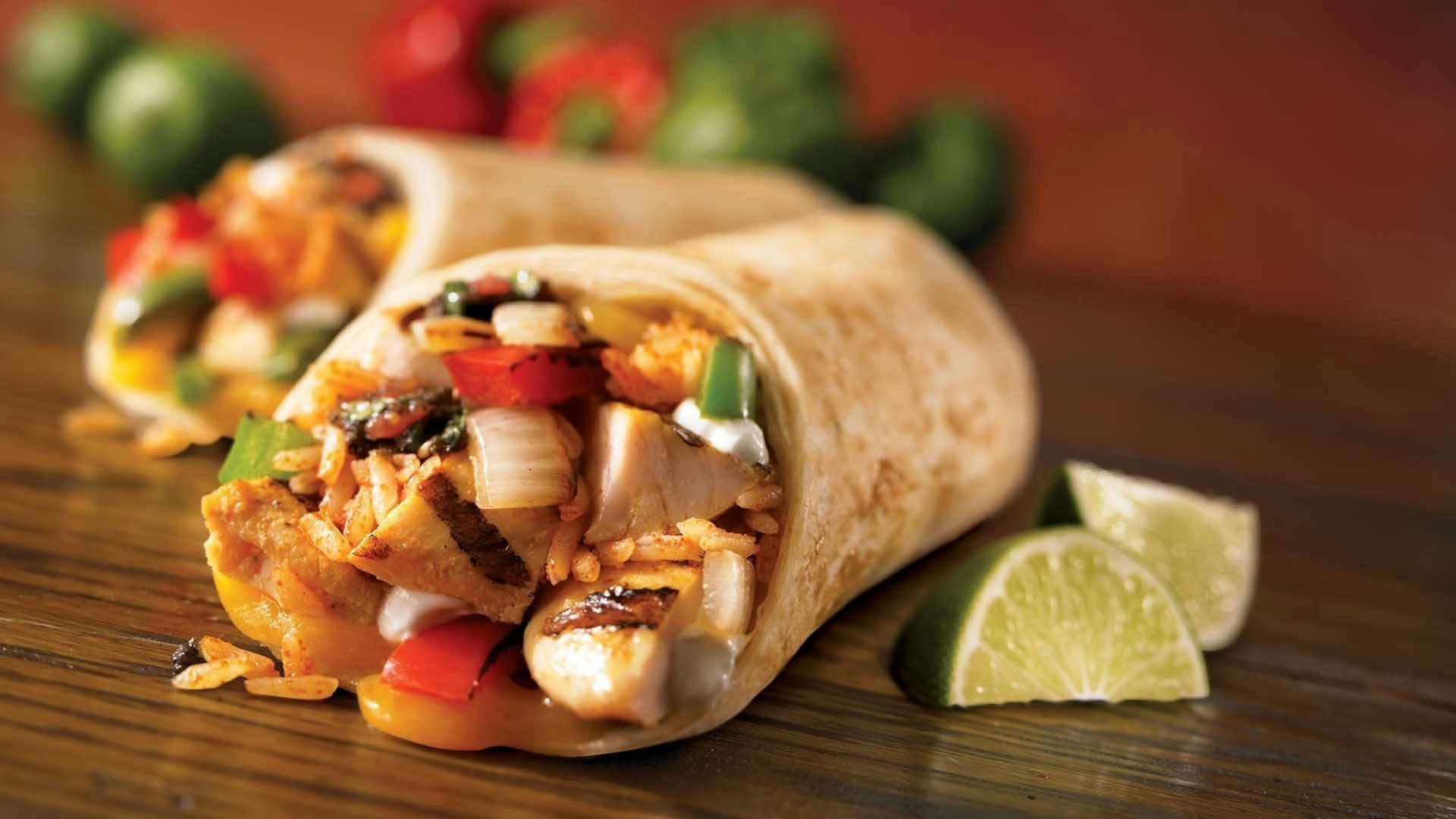 Two chicken and rice burritos on a wooden surface with lime wedges and peppers in the background.
