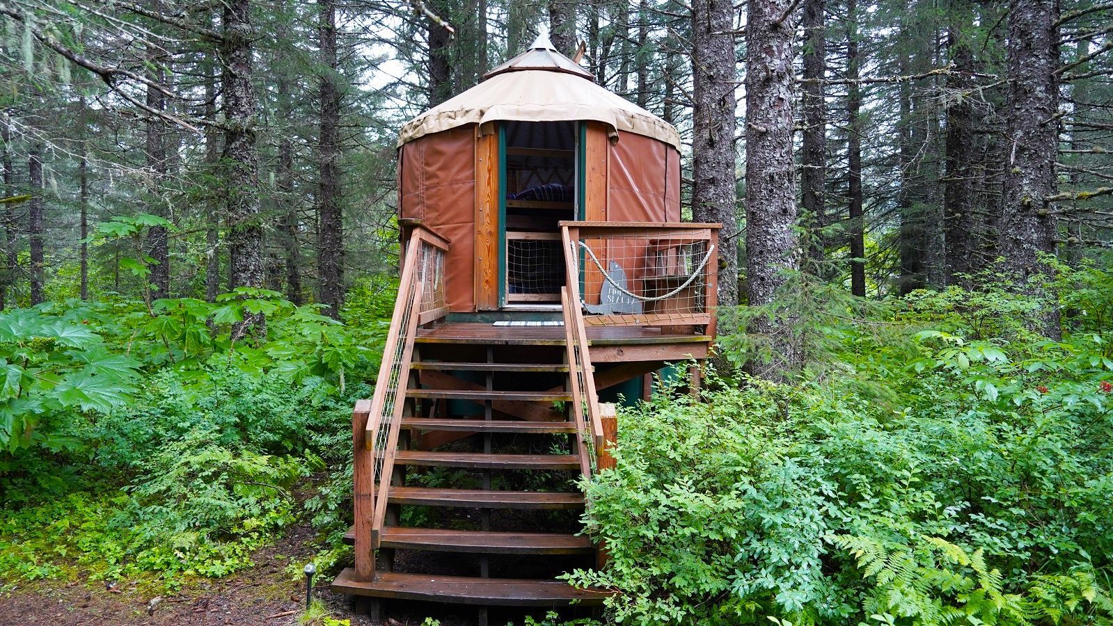 Wooden yurt with a deck, accessed by stairs, surrounded by green foliage in a forest.