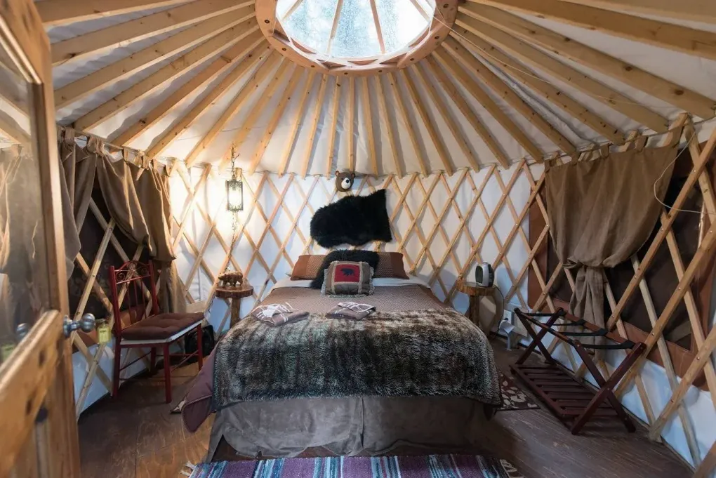 Interior of a yurt with bed, chair, windows, and skylight. Earth-tone decor with brown wood and tan fabric.