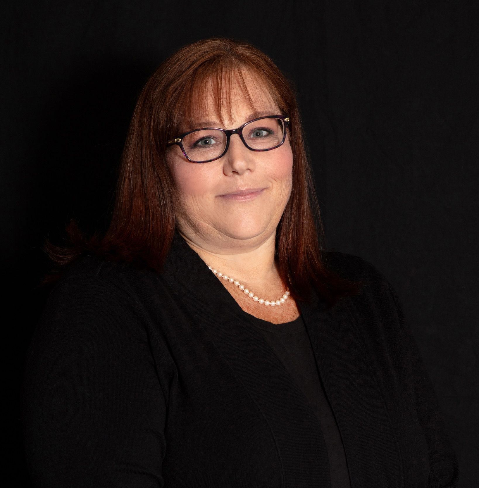 A woman wearing glasses and a pearl necklace is standing in front of a black background.