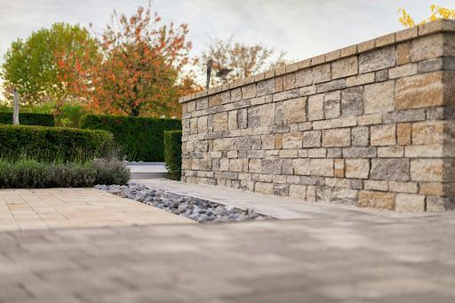 A stone wall sits beside a paved patio and gravel path, bordered by manicured hedges and autumn-toned trees.