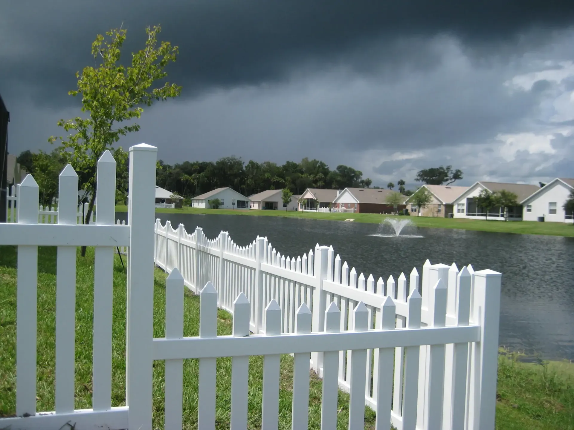A white picket fence surrounds a lake with houses in the background