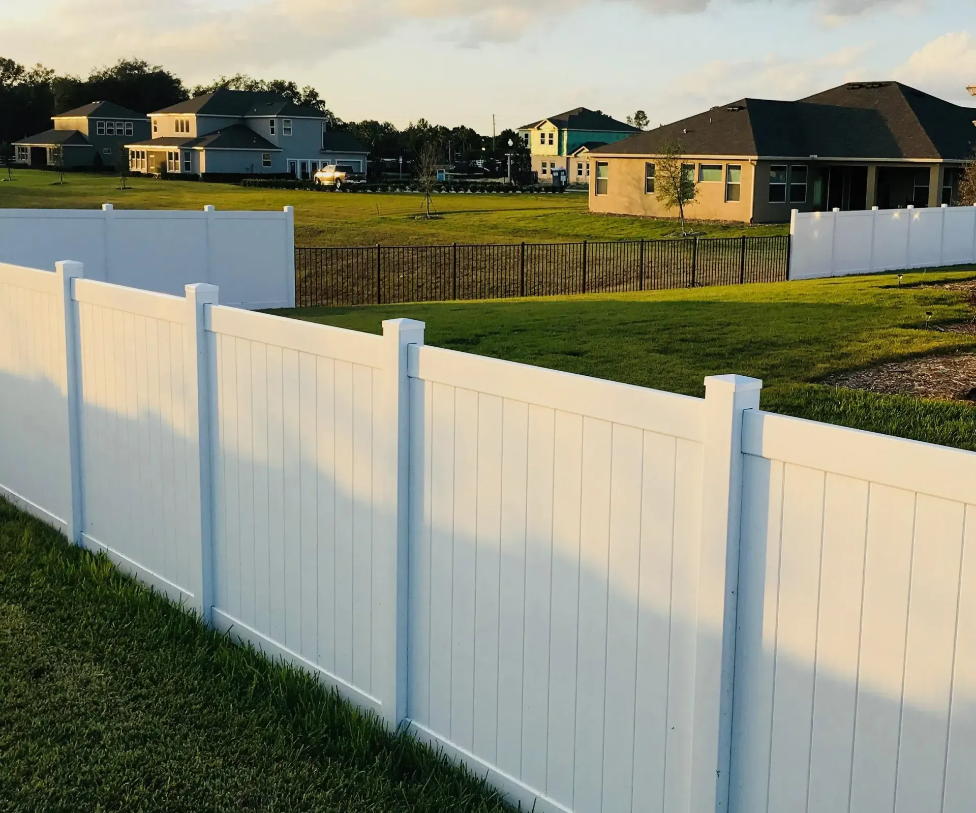 A white fence surrounds a grassy field with houses in the background.