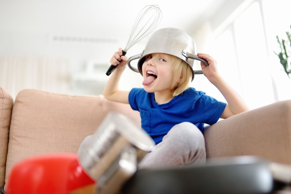 A child sits on a couch wearing a metal pot as a hat, sticking their tongue out while holding a whisk.