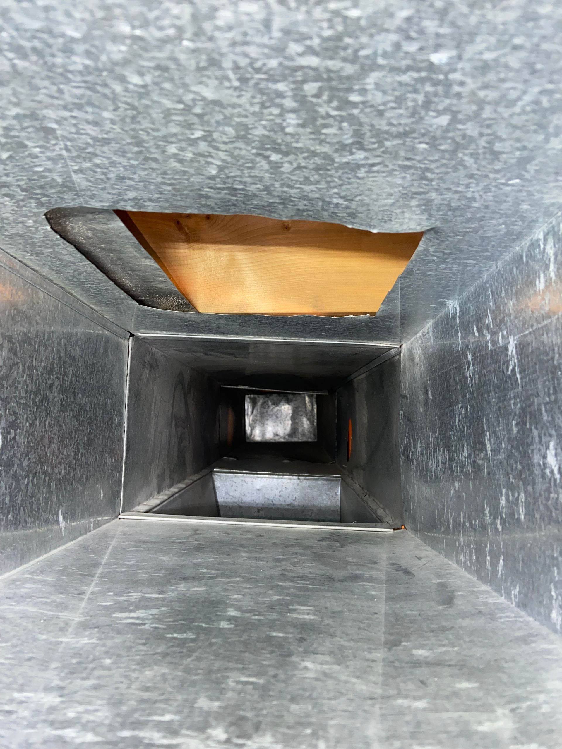 Interior of a metal air duct with a rectangular opening and a wooden panel visible near the top opening.