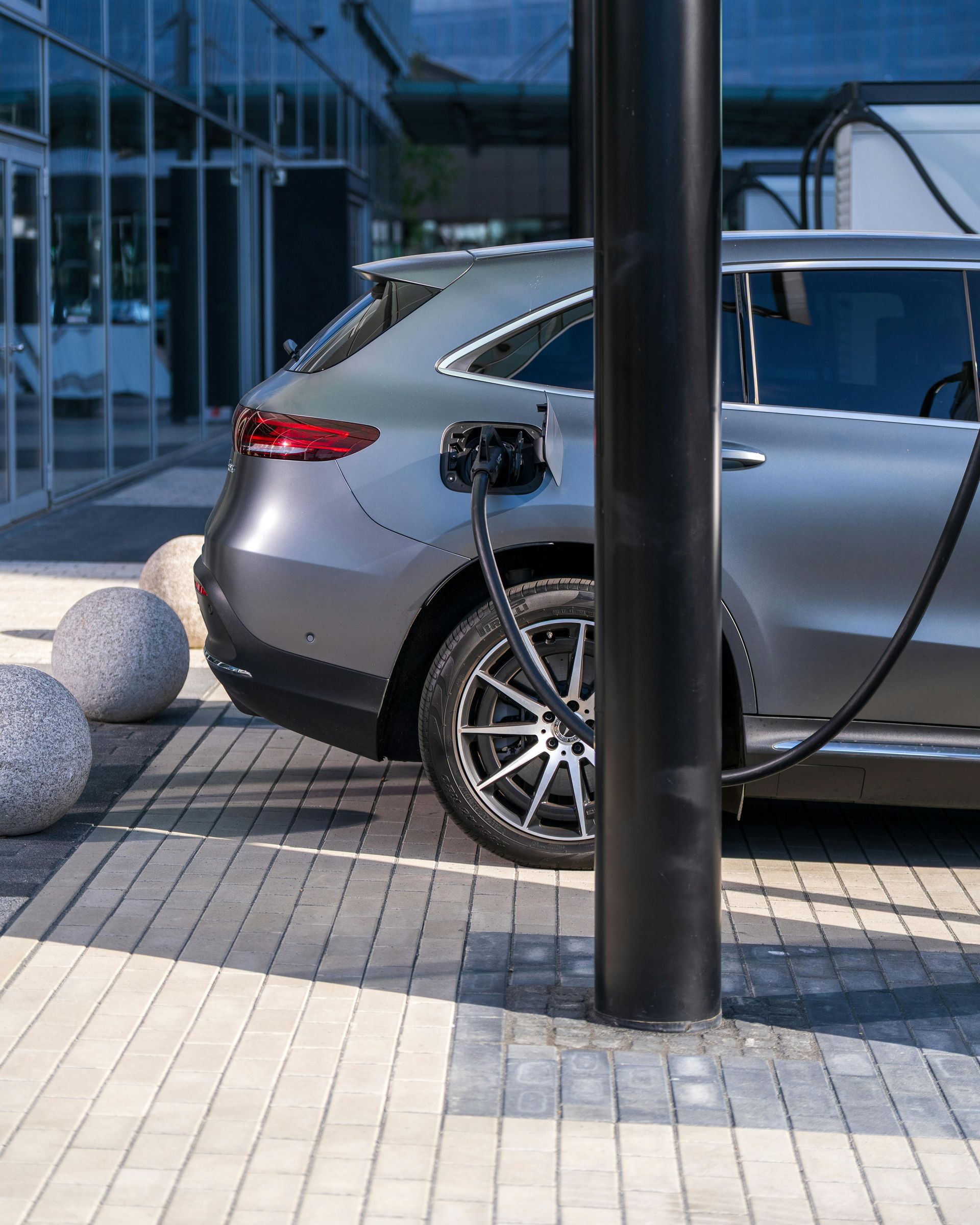 Silver SUV parked beside a black pole on a tiled sidewalk near a glass building.