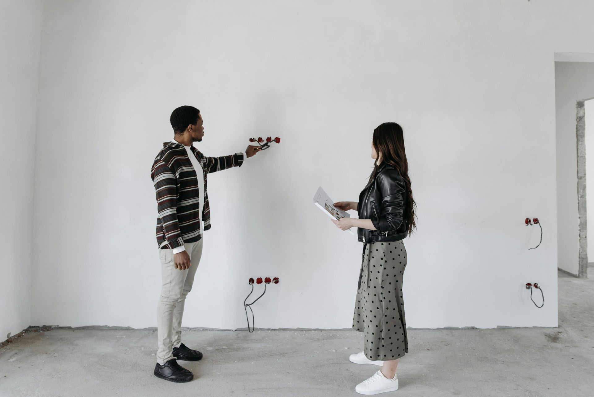 Two people discussing plans in an empty white room with exposed wall outlets and unfinished floor.