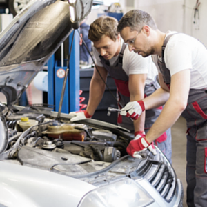 Two mechanics, in gray and red overalls, working on a car engine in a repair shop.