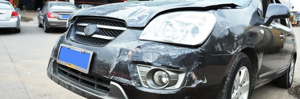 A black car with front-end damage; appears to have been in a collision. Other cars are parked nearby.