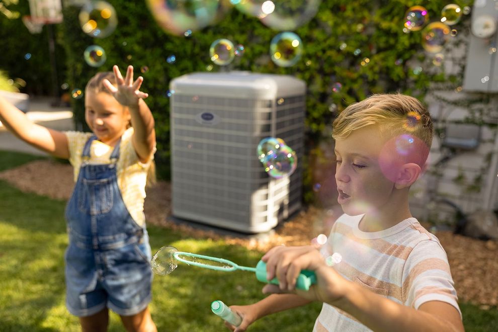 Children blowing bubbles in a sunny yard near an air conditioner unit