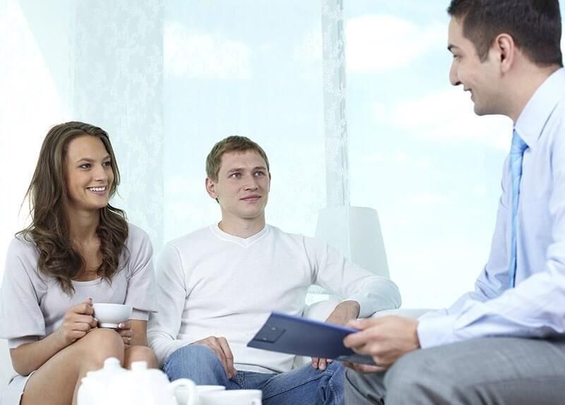 Un hombre y una mujer están sentados en un sofá hablando con un hombre que sostiene un portapapeles.