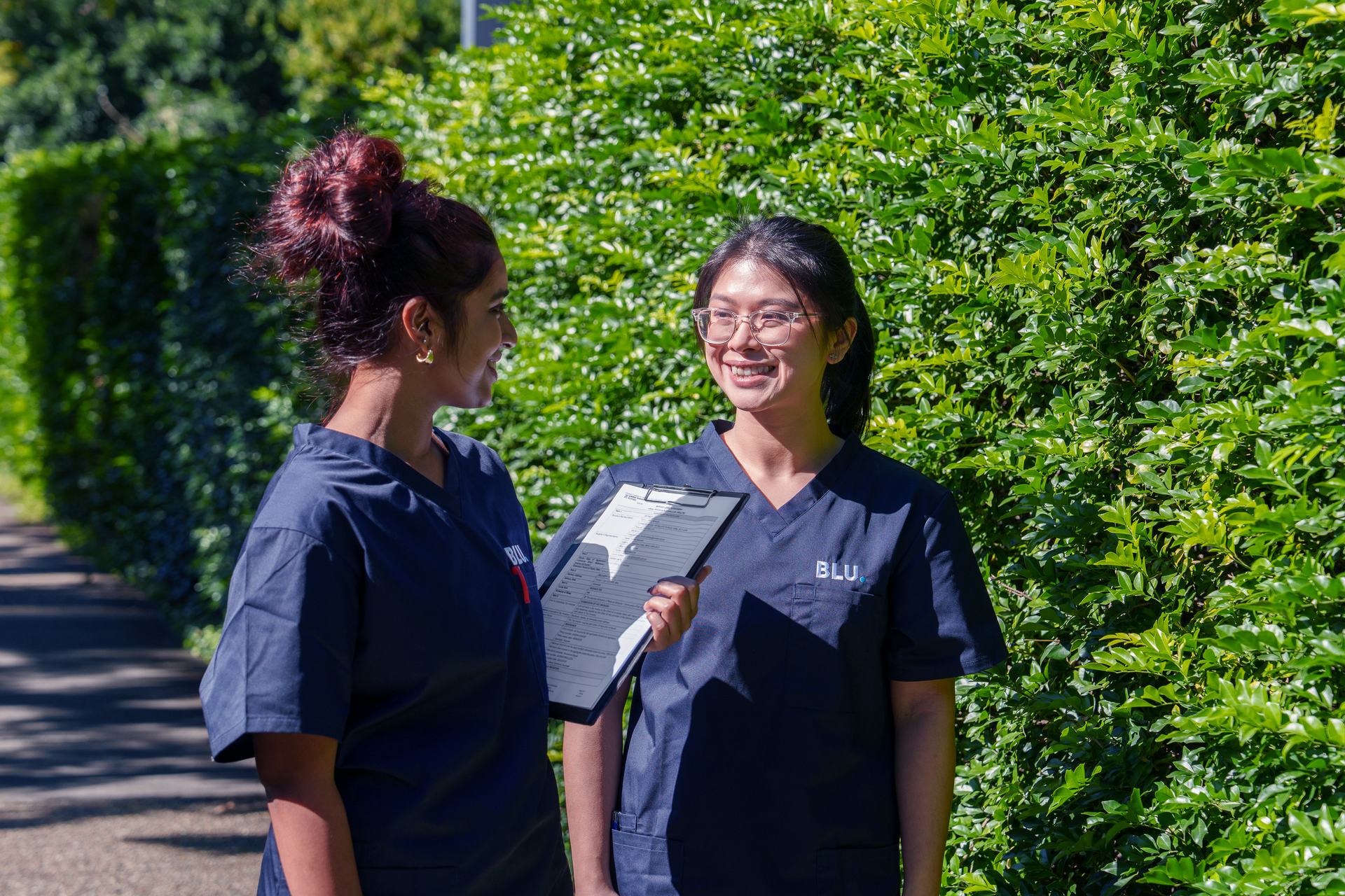 Two nurses are standing next to each other in front of a hedge.