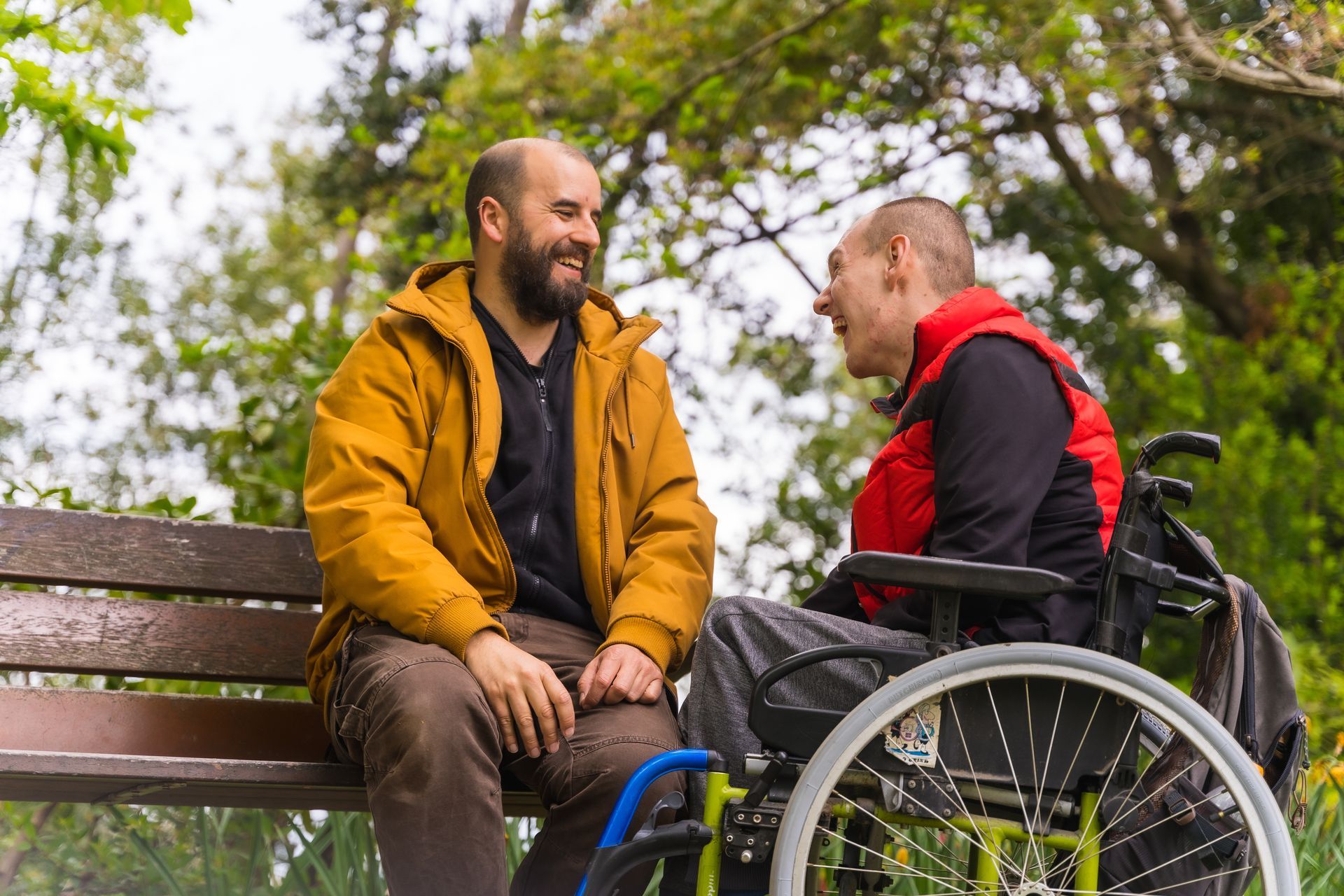 A man in a wheelchair is sitting on a park bench talking to another man.